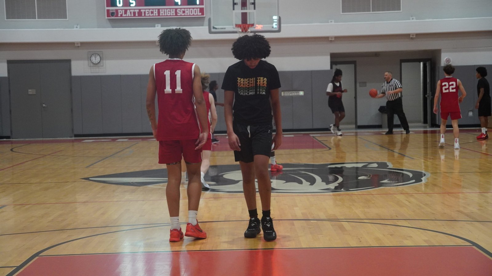 Two young men, one wearing a red #11 jersey and the other a black Tai Chi Tea shirt, stand on a basketball court at Platt Tech High School. Other players and a referee are visible in the background.